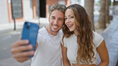 Gülümseyen bir çift güneşli bir şehir sokağında birlikte selfie çeker. Aşkı, birlikteliği ve modern şehir yaşamını özetlerler..