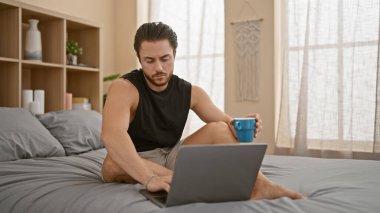 Young hispanic man using laptop drinking coffee at bedroom