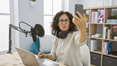 Middle-aged woman live-streaming in a modern radio studio with a microphone and laptop. clipart