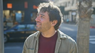Genç bir İspanyol adam, güneşli bir şehir caddesinde dururken nazikçe gülümsüyor, şehir yaşamını bir tarz dokunuşuyla özetliyor..