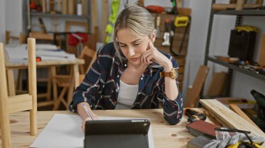 Odaklanmış genç kadın marangozluk atölyesinde tablet üzerine notlar alıyor