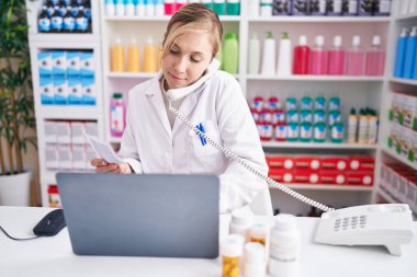 Young blonde woman pharmacist talking on telephone using laptop at pharmacy