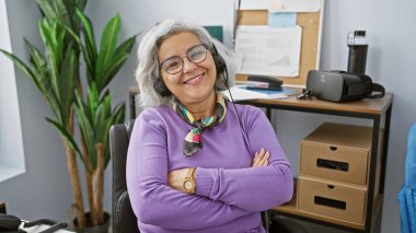 Gülümseyen, gri saçlı, kulaklık takan, kollarını kavuşturan, kendine güvenen olgun bir kadın..