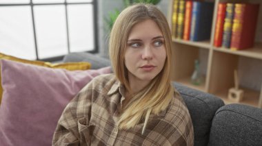 Düşünceli genç bir kadın, yumuşak yastıklar ve arka planda bir kitaplıkla çevrili, kapalı bir kanepede dinlenir..