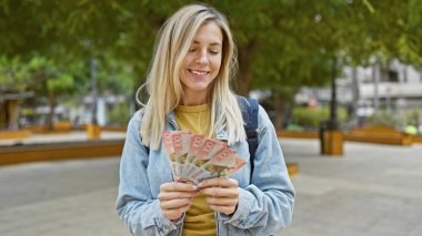 Gülümseyen sarışın kadın parkta yeni Zelanda dolarları sayıyor..