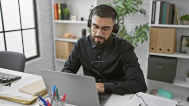 Kulaklık takan yakışıklı sakallı adam modern ofis ortamında dizüstü bilgisayarla çalışıyor..