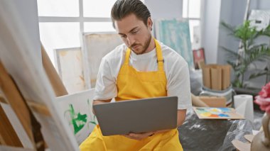 Genç İspanyol ressam, sanat stüdyosunda ciddi yüzlü dizüstü bilgisayar kullanıyor.