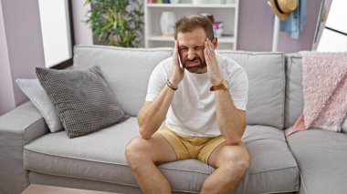 Evde baş ağrısından muzdarip, gri saçlı olgun bir adam, kapalı bir kanepede oturuyor..