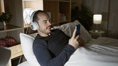 Kulaklıklı İspanyol bir adam gece yatak odasında akıllı telefon kullanıyor.