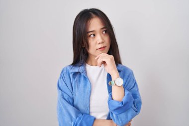 Beyaz arka planda, elleri çenesinde soru sormayı düşünen genç Çinli bir kadın. Düşünceli bir ifade. Gülümseyen ve düşünceli bir yüz. şüphe kavramı. 