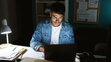 Kot ceketli, odaklanmış bir İspanyol gece geç saatlere kadar loş bir ofiste dizüstü bilgisayarda çalışır..