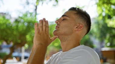 Beyaz tişörtlü, yeşil bir parkın içinde huzur içinde dua eden İspanyol bir adam.