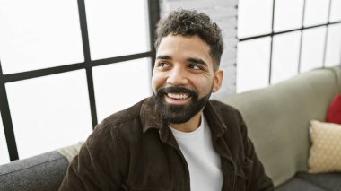 Gülümseyen, sakallı genç bir adam modern bir oturma odasında rahatça oturuyor, evde rahatlama tasvir ediyor..