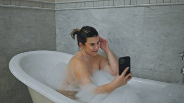 Gülümseyen bir kadın fayanslı bir banyoda akıllı telefonunu karıştırırken köpüklü bir küvetin tadını çıkarır..