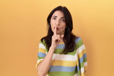 Middle age hispanic woman standing over yellow background asking to be quiet with finger on lips. silence and secret concept. 