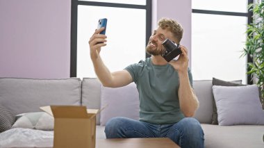Young, handsome man with a beard taking a selfie indoors while unpacking a box in a modern living room. clipart