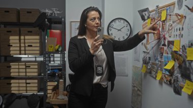 Yetişkin kadın polis dedektifi kanıt panosunu incelerken kapalı ofis ortamında sesli mesaj kaydediyor..