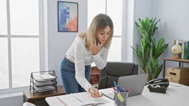 Modern bir ofiste çalışan, notlar yazan ve telefonla konuşan beyaz bir kadın..