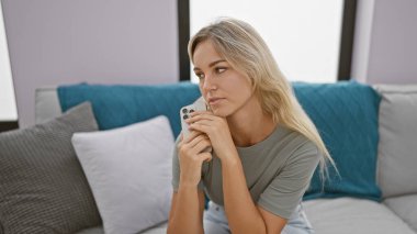 Düşünceli, genç, sarışın bir kadın evinde rahat, rahat, modern bir oturma odasında akıllı bir telefon tutuyor..