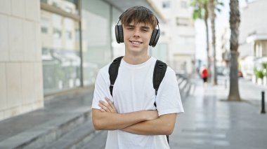 Kulaklıklı ve sırt çantalı genç sarışın bir adam güneşli bir şehir caddesinde gülümsüyor..