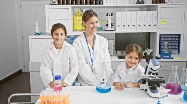 Laboratuvar önlüğü giyen gülümseyen bir kadın ve iki kız mikroskop ve beher ile laboratuvar bankında oturuyorlar..