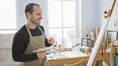 Parlak bir sanat stüdyosunda, sakallı, gülümseyen kel bir adam tuvale resim çiziyor..
