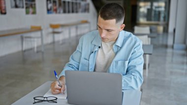 Yakışıklı İspanyol genç adam üniversite kütüphanesinde dizüstü bilgisayar ve defterle çalışıyor..