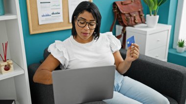 A middle-aged hispanic woman examines a credit card while sitting with a laptop in a modern office interior. clipart