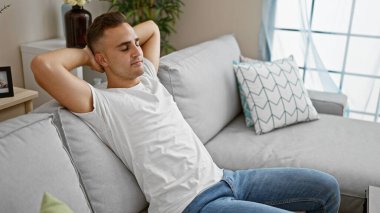 Rahat genç bir adam, parlak, modern bir oturma odasında, bir kanepede uzanıyor, gündelik rahatlığı somutlaştırıyor..