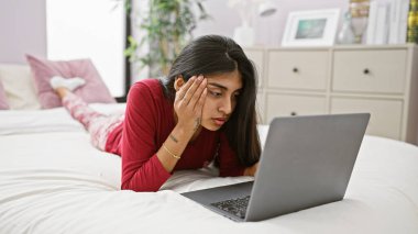 Kırmızılı odaklı Hintli bir kadın rahat bir yatak odasında beyaz bir yatakta yatarken dizüstü bilgisayarla çalışıyor..