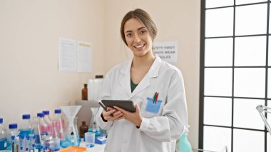 Laboratuvarında teçhizat ve deney şişeleriyle çevrili tabletle gülümseyen kadın bilim adamı.