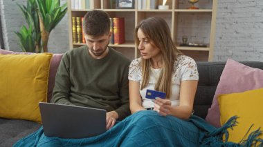 Bir adam ve bir kadın birlikte kanepede oturuyor, dizüstü bilgisayar kullanıyor ve evde kredi kartı tutuyor..