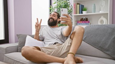 Modern oturma odasındaki koltukta selfie çeken sakallı adam, gülümsüyor, barış işareti yapıyor, rahat, kapalı, İspanyol, yetişkin, erkek, yakışıklı, eğlenceli.