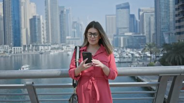 Brunette woman in red uses smartphone against dubai marina skyline backdrop, embodying luxury, travel, and modernity. clipart