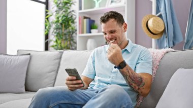 İspanyol adam rahat bir oturma odasında kanepede otururken telefona gülümsüyor.