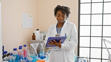 Afrika kökenli Amerikalı kadın bilim adamı bir laboratuarda cam ve ekipmanla notları inceliyor.