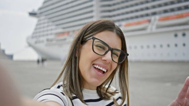 Gözlüklü, gülümseyen genç bir kadın deniz yolculuğu sırasında lüks bir yolcu gemisinin önünde selfie çekiyor..