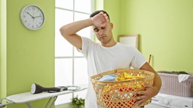 Parlak yeşil bir odada çamaşır sepetini tutan yorgun genç bir adam, bunalım ve yorgunluk ifade ediyor..