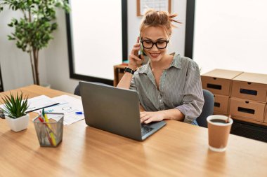 Kafkasyalı genç bir iş adamı bilgisayarını kullanarak ofisteki akıllı telefondan konuşuyor.