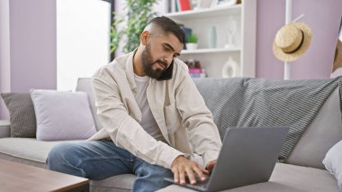 Sakallı yakışıklı bir adam rahat bir oturma odasında dizüstü bilgisayarı ve telefonu olan birden fazla iş yapıyor..
