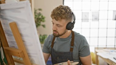 Kulaklıklı, parlak bir sanat stüdyosundaki tuvale resim yapan odaklanmış genç bir adam.
