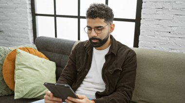 Sakallı, gözlüklü genç bir İspanyol evdeki koltukta oturup tablete odaklanmış..