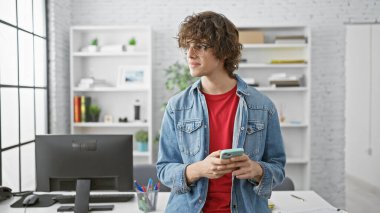 Modern ofis ortamında akıllı telefon kullanan kıvırcık saçlı ve gözlüklü sıradan bir genç..