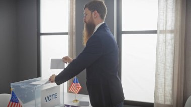 Amerikan seçimlerinde oy kullanan takım elbiseli sakallı bir adam, kapalı alanda bayraklarla.