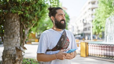 Sakallı, çörek gibi saçı olan gülümseyen İspanyol adam parkta akıllı telefondan mesaj atıyor.