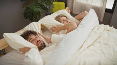Bir adam ve bir kadın yatak odasında yatarken kulaklarını tıkarlar, gürültüden rahatsız olmuş görünürler.