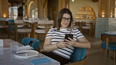 Modern restoranda akıllı telefon kullanan genç bir kadın. Teknoloji, yaşam tarzı ve yemek atmosferi sergileniyor.