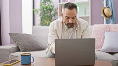 Gri sakallı olgun bir İspanyol adam rahat bir oturma odasında dizüstü bilgisayarla çalışıyor ve evde üretkenlik hissi veriyor..