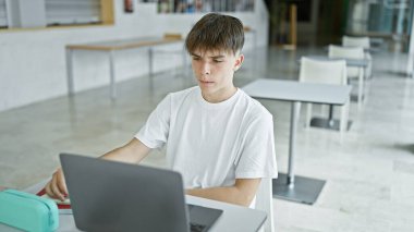 Genç sarışın erkek öğrenci modern bir üniversite kütüphanesinde dizüstü bilgisayarla çalışıyor.