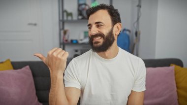 Gülümseyen sakallı adam rahat, aydınlık bir oturma odasını işaret ediyor..